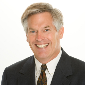 Person looking directly forward, smiling, wearing a black blazer, white collard shirt, and maroon tie.