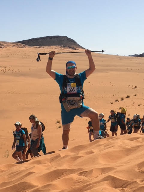 Athlete at top of a sand dune with arms raised triumphantly over head.