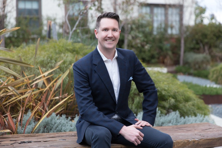 Photograph of Scott Malcolm, sitting outside, smiling and looking directly forward, wearing a dark business suit with white collared shirt