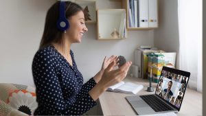 Adult wearing headphones, smiling, gesturing with her hands, looking at laptop which displays attendees of online video call.