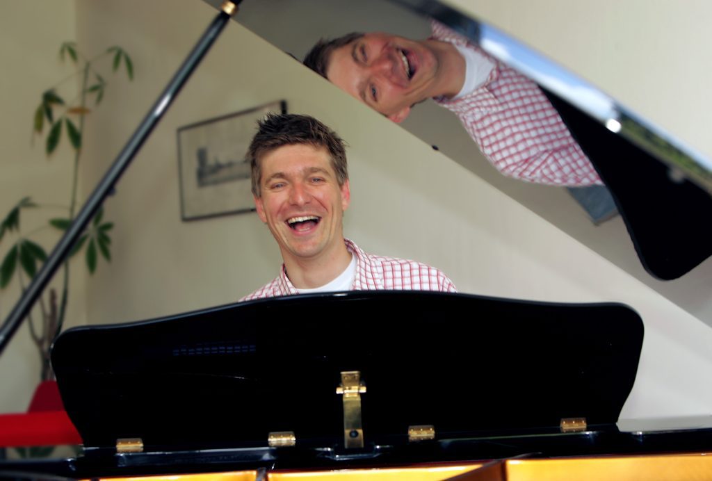 Man laughing at a grand piano with his reflection in the lid.