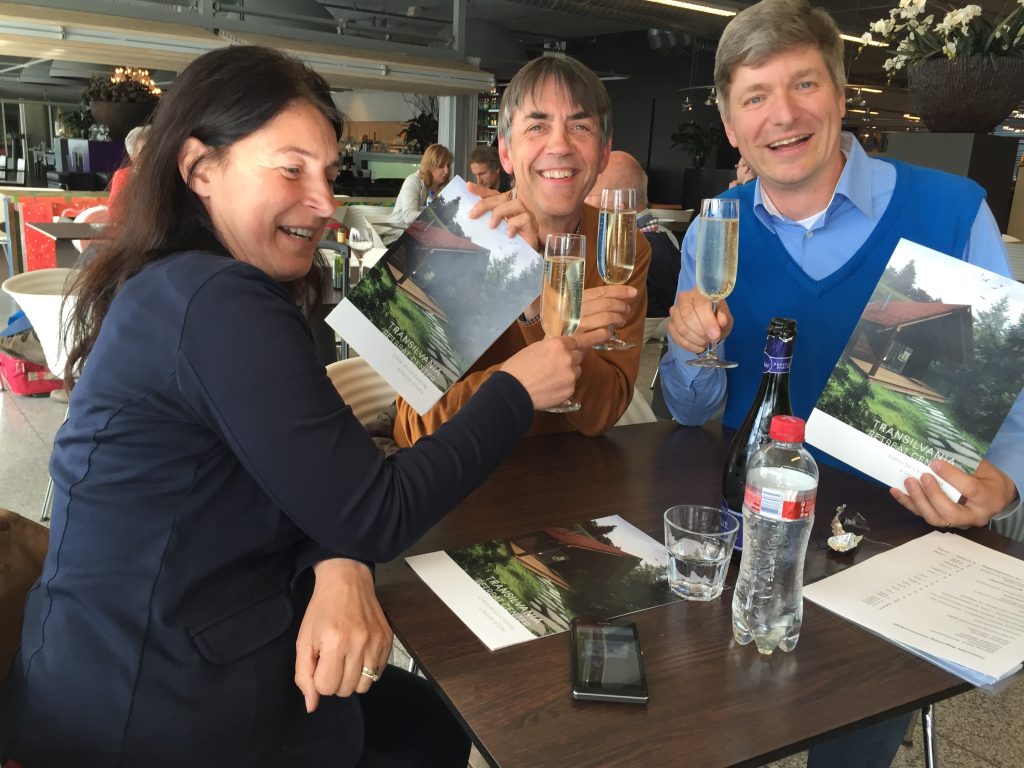 Three colleagues smiling and toasting champagne while holding flyers.