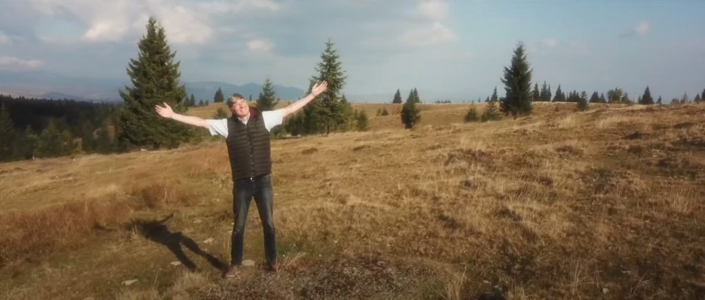 Man in a white tee shirt, black vest, and jean with his arms open wide staring into the sun with a smile surrounded by a grassy landscape.