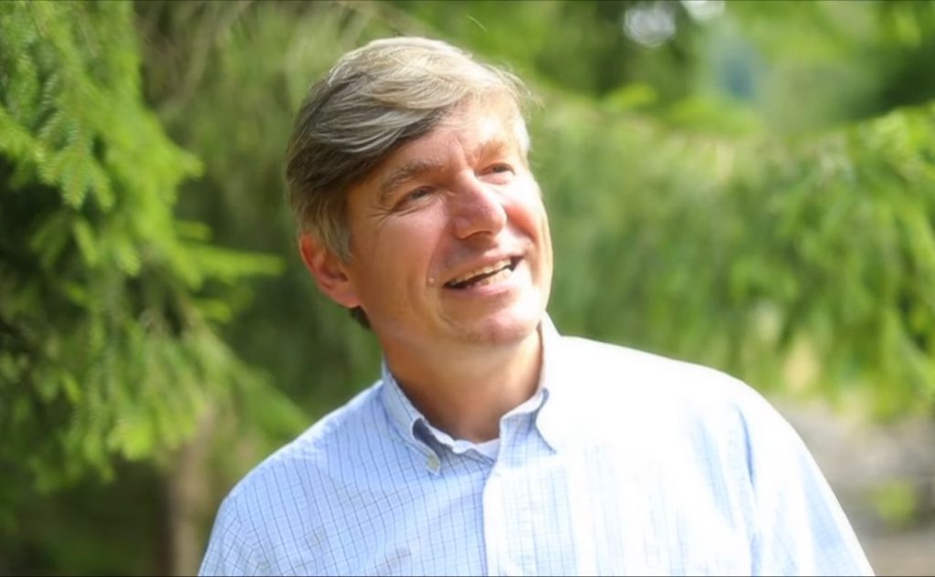 Man in a collared shirt smiling in front of an evergreen tree.